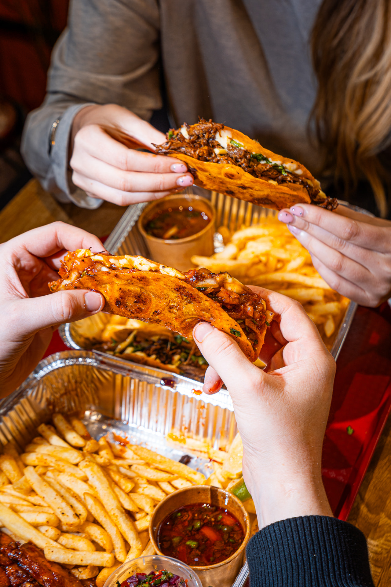 Hands holding birria tacos over a sharing tray of fries